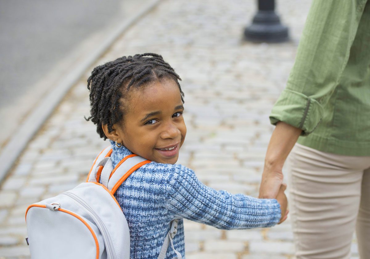 young boy with backpack on holding his mother's hand, looking back at camera and smiling