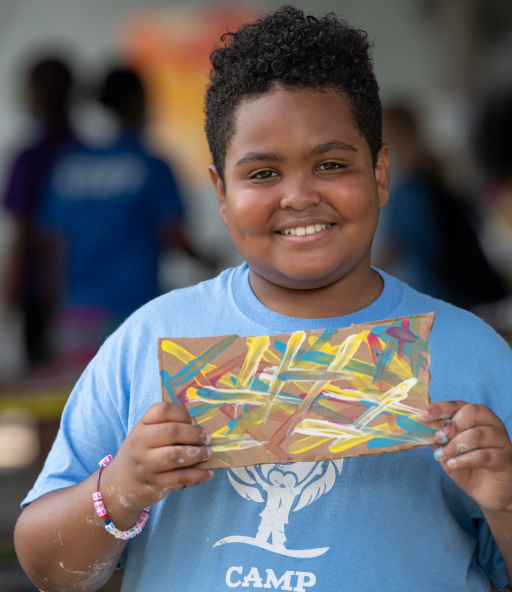 young camper wearing light blue Camp Harbor View t-shirt, smiling and holding his artwork, on the island