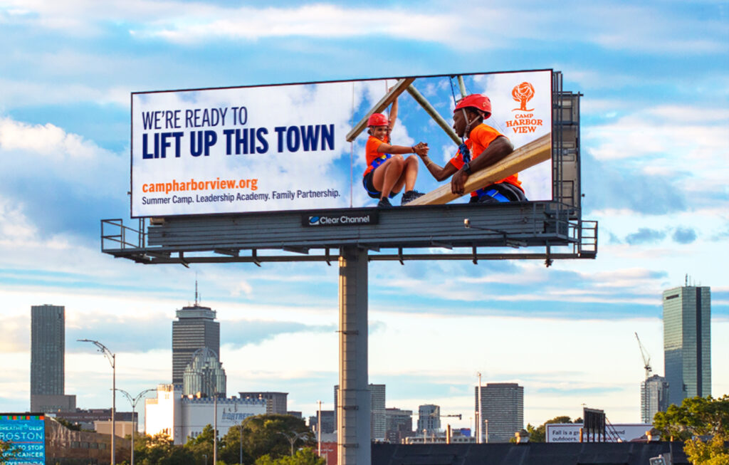 Photo of a digital Camp Harbor View billboard on route 93 in Boston that says "WE'RE READY TO LIFT UP THIS TOWN" with an image of one teen helping another on the ropes course on the summer camp island.