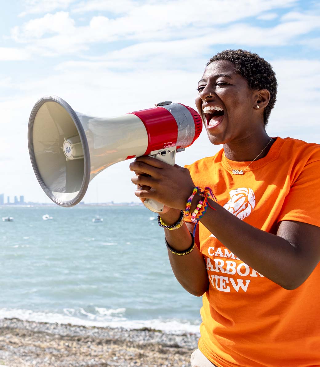 Camper in orange Camp Harbor View t-shirt cheering into a bullhorn, standing on Long Island with a view of the Boston skyline