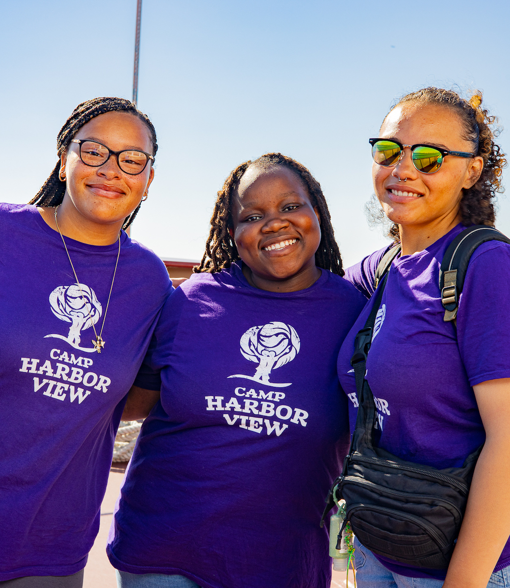 Three teenagers in purple Camp Harbor View shirts smiling at the camera on a sunny day.