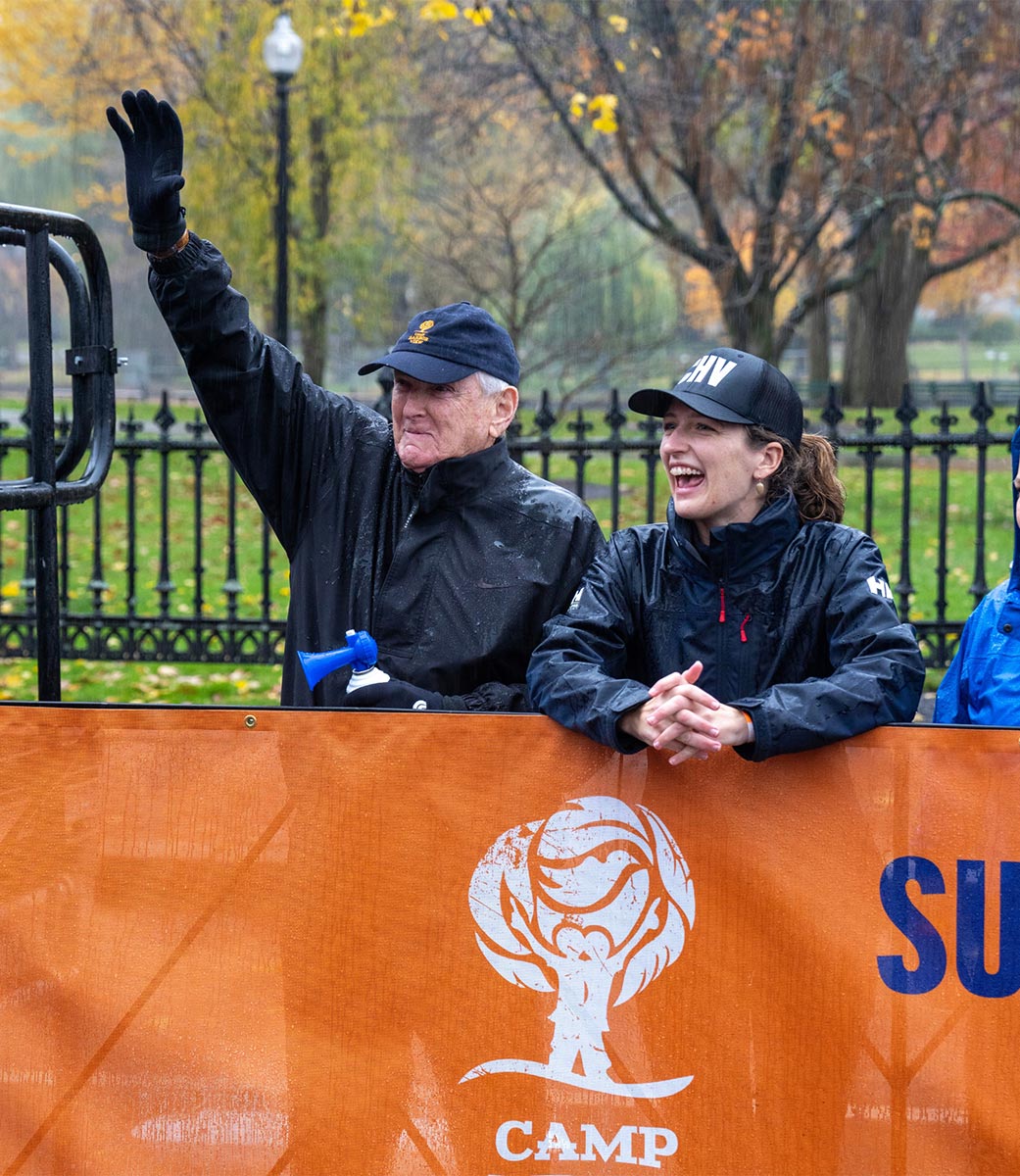 Jack Connors and friend waving from the sidelines at Camp Harbor View Road Race on the Boston Common, orange banner