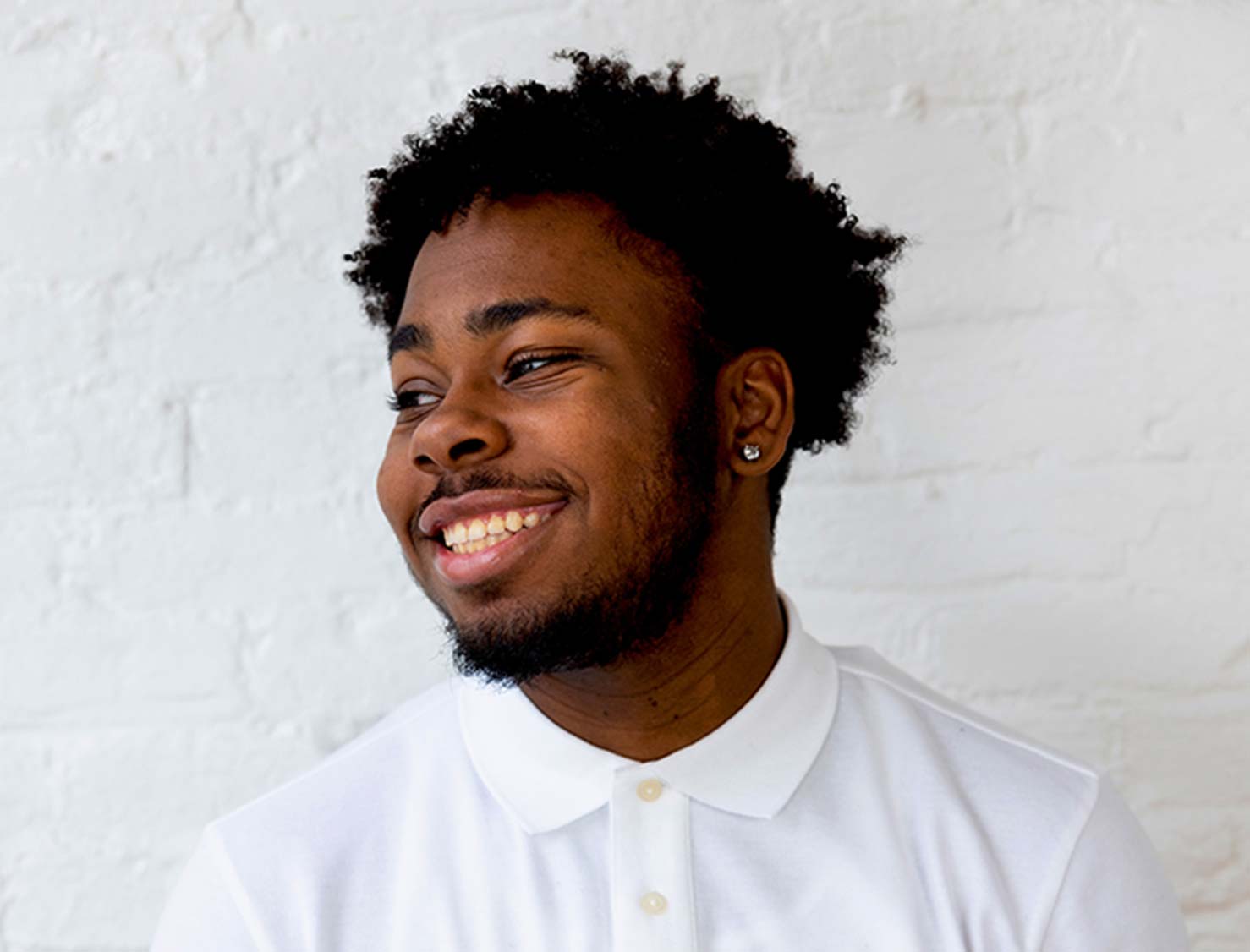 A young man smiles while looking to his left. He is wearing a white buttoned polo shirt and diamond stud earrings. The background is a white brick wall.