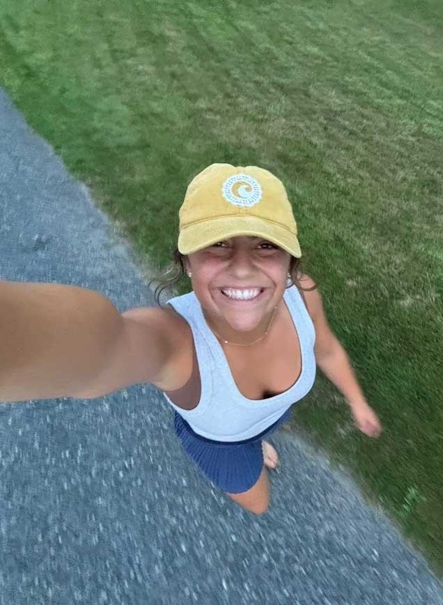 Woman smiling while walking outdoors, wearing a yellow cap and athletic tank top.