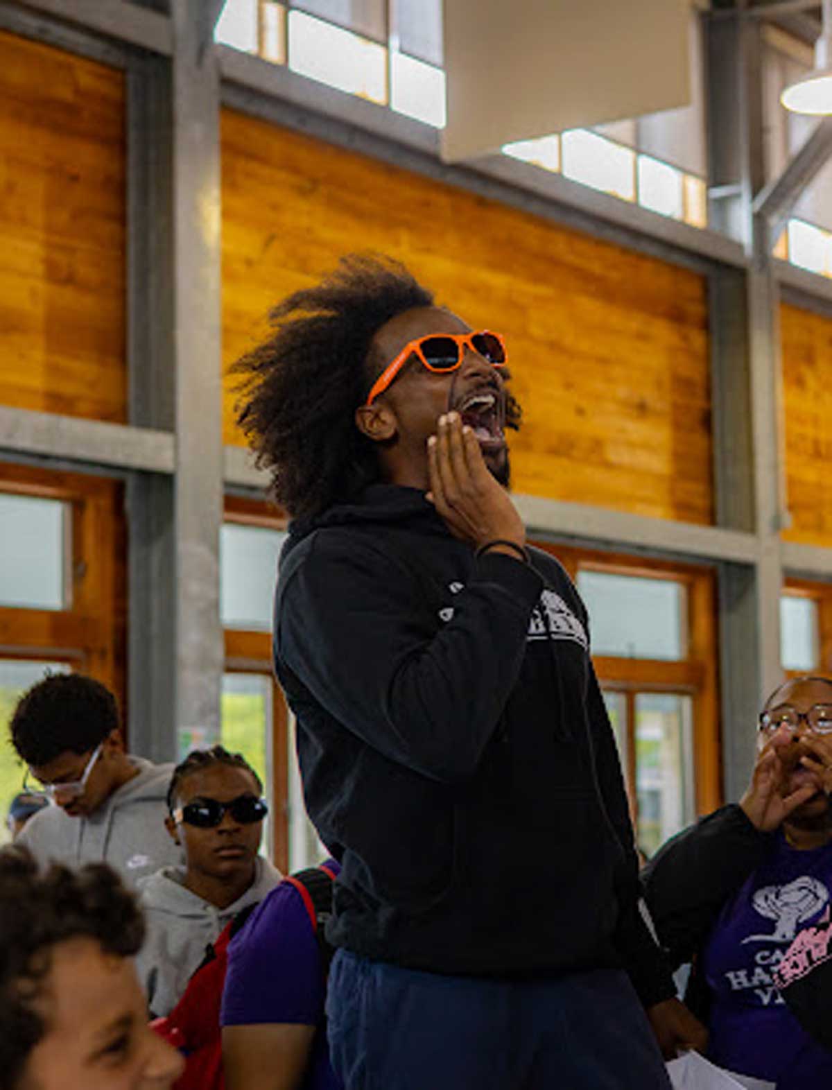 Man cheering enthusiastically indoors, wearing bright orange sunglasses and a black hoodie, surrounded by a crowd.