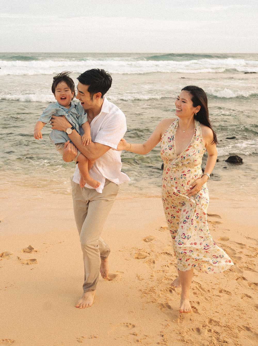 Family walking barefoot along a sandy beach, smiling as they hold a young child near the ocean waves.