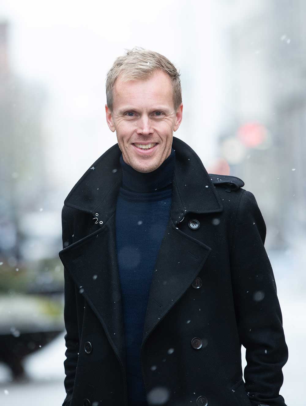 Man standing outdoors in light snowfall, wearing a dark coat and smiling toward the camera.