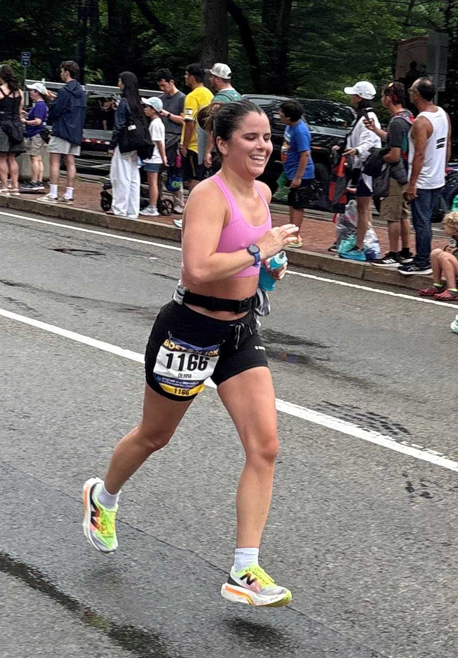 Woman running in a road race wearing a pink sports bra and race bib, with spectators along the route.