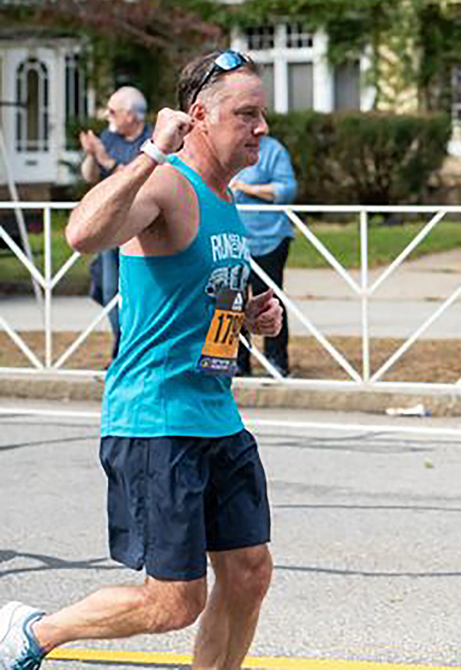 Man running in a road race wearing a teal tank top and race bib, raising his fist while spectators cheer behind him.