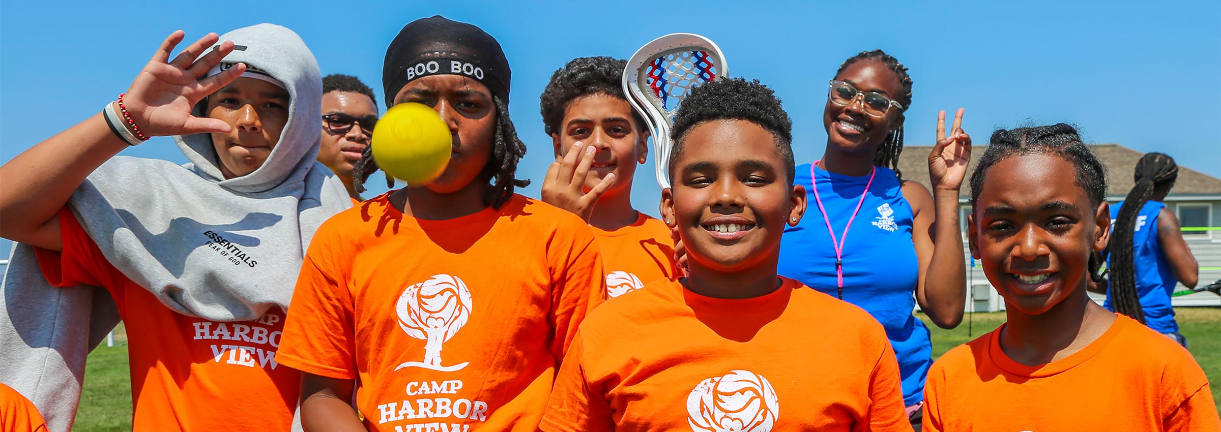 group of campers smiling and having fun wearing orange Camp Harbor View t-shirts, with a blue sky behind them